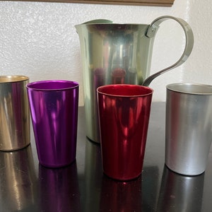 May include: A collection of metallic drinkware, including a silver pitcher with a curved handle and four colorful tumblers. The tumblers are gold, purple, red, and silver. The pitcher and tumblers are arranged on a dark surface.