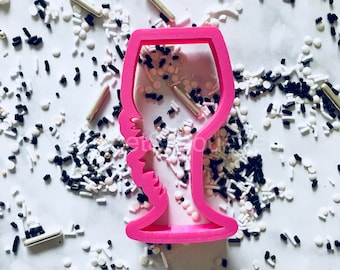 May include: Pink plastic wine glass cookie cutter. The cutter is on a white surface with black and white sprinkles.