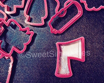 May include: Pink plastic cookie cutters in various shapes, including a tree, a star, a bell, a boot, and an axe. The cutters are on a sparkly, glittery surface.