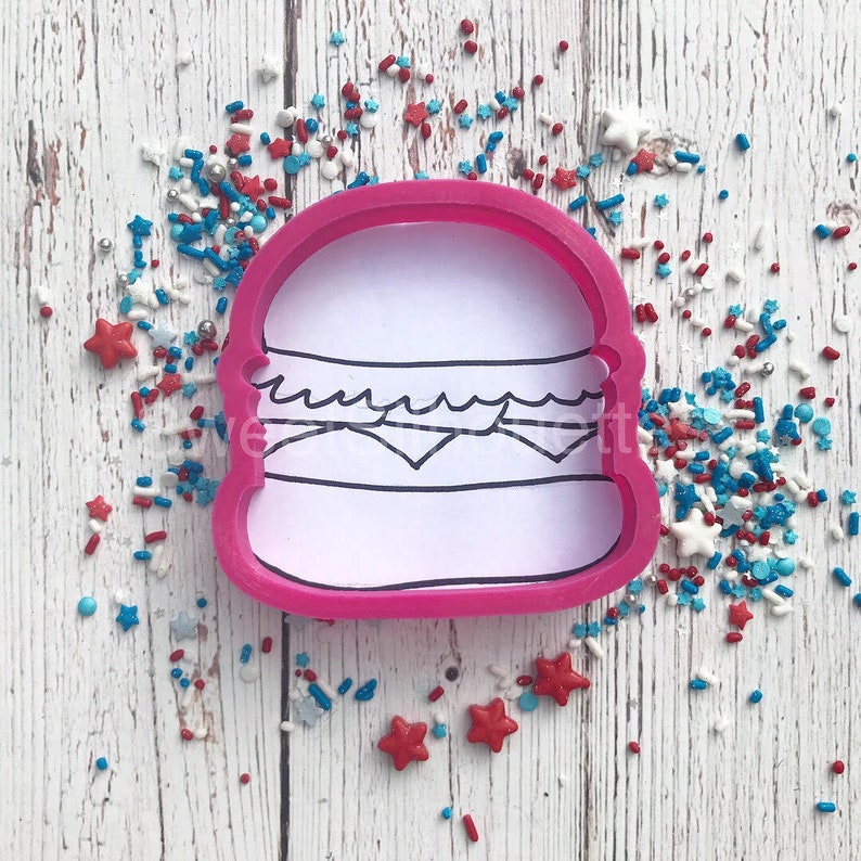Pode incluir: Um cortador de biscoitos de pl&aacute;stico rosa em forma de hamb&uacute;rguer, com interior branco e contorno preto detalhando os componentes do hamb&uacute;rguer. Polvilhos e decora&ccedil;&otilde;es em forma de estrela vermelhas, brancas e azuis est&atilde;o espalhados ao redor.