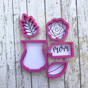 May include: Pink plastic cookie cutters in the shapes of a vase, a rose, a leaf, and a tag that says "mom".