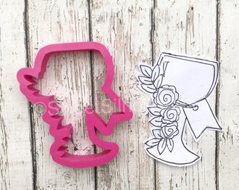 May include: Pink plastic cookie cutter shaped like a bride with a bouquet of flowers. A black and white outline of the same design is shown on a white background.