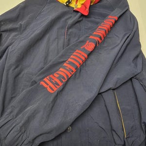 May include: A navy blue jacket with a red and yellow collar. The sleeve features the name "Tommy Hilfiger" in red lettering. The jacket has a zipper closure and button details. A classic, sporty outerwear piece.