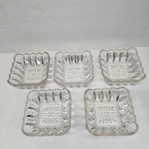 May include: Five clear glass square dishes with a decorative floral pattern in the center. The edges of the dishes have a scalloped design. The dishes are arranged on a white surface.