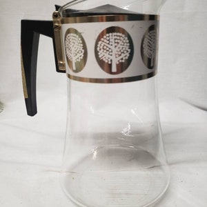 May include: A clear glass pitcher with a black handle and a gold band around the top. The band has a white design of three stylized trees.
