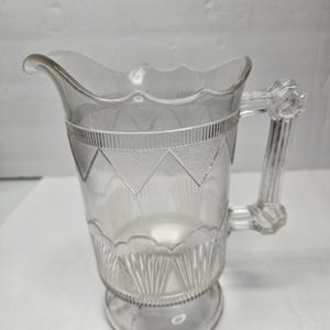 May include: Clear glass pitcher with a decorative handle and spout. The pitcher features etched geometric patterns around the body and a scalloped rim. The handle has a detailed design. The pitcher is likely for serving beverages.