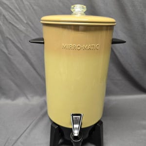 May include: A vintage Mirro-Matic percolator with a cylindrical yellow body, a matching lid with a clear glass knob, and black side handles. The base is black with three legs and a central black spigot for dispensing liquid from the container.