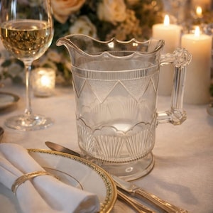 May include: A clear glass pitcher with a decorative pattern, a wine glass, and a plate with a gold rim are set on a white tablecloth. The scene is illuminated by candlelight and a floral arrangement.
