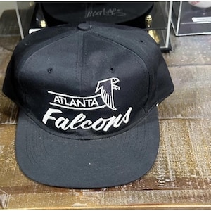 May include: Black Atlanta Falcons baseball cap with a flat brim. The cap features the team name and logo in white embroidery on the front. The text "ATLANTA" is above the logo, and "Falcons" is below.