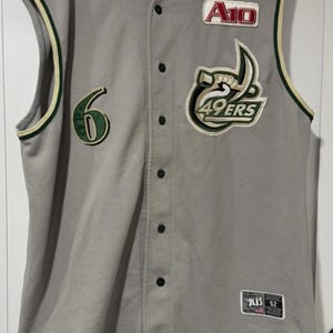 Op de afbeelding: Grijs honkbalshirt met groene en witte accenten. Voorzien van nummer 6, een patch met "A10" en een "49ERS" logo. Het shirt heeft een knoopsluiting en een label met "AIS 52".