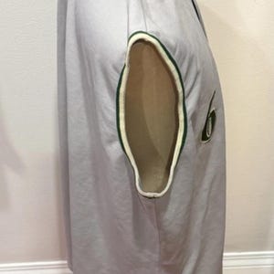 Op de afbeelding: Een lichtgrijs honkbalshirt met groen-witte randen op de mouwopeningen. Het shirt heeft een nummer 6 patch op de mouw. Het shirt wordt op een mannequin getoond.