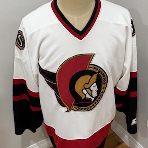 May include: White hockey jersey with a V-neck and the Ottawa Senators logo on the front. The jersey has red and black accents on the sleeves, collar, and hem. The logo features a gladiator's head.