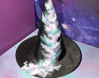 Puede incluir: Un sombrero de bruja negro con un ala ancha y una corona alta y puntiaguda. El sombrero está pintado con un diseño de galaxia con nubes giratorias de color rosa, azul y blanco sobre un fondo negro.