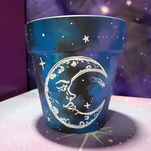May include: A tapered cylindrical plant pot painted with a dark blue and black galaxy-themed background. The exterior features a hand-painted white crescent moon with a human-like face and small white stars scattered across the surface.