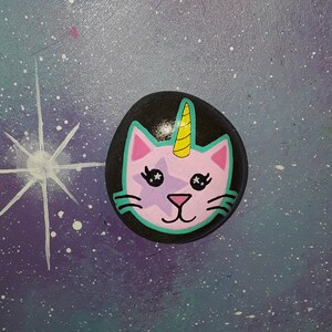 Puede incluir: Una piedra negra pintada a mano con una cara de gato rosa, una estrella morada, ojos en forma de estrella y un cuerno de unicornio amarillo. La cara del gato está delineada en verde azulado. El fondo es un cielo estrellado.