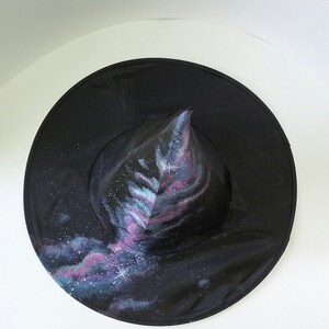 May include: A black witch hat with a pointed crown painted with a galaxy design. The hat features a swirling pattern of blue, purple, and pink colors with white stars.