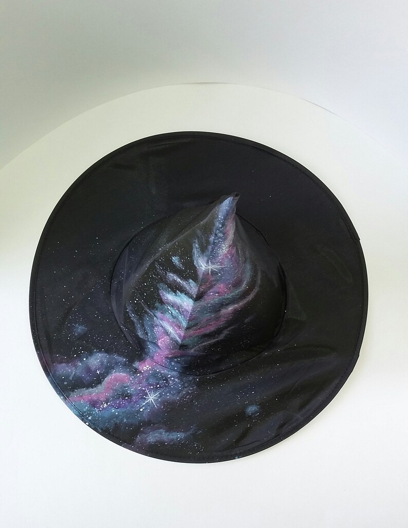 May include: A black witch hat with a pointed crown painted with a galaxy design. The hat features a swirling pattern of blue, purple, and pink colors with white stars.