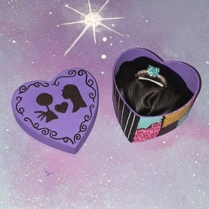 May include: A silver-toned ring with a heart-shaped blue stone sits inside a heart-shaped box. The box lid is purple with black silhouettes of two figures and a heart. The box base features a patchwork pattern of black, pink, and yellow.