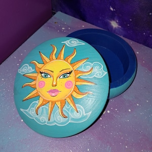 Könnte beinhalten: Eine runde Holzkiste mit einem blauen Deckel. Der Deckel ist mit einer gelben Sonne mit einem lächelnden Gesicht, rosa Wangen und blauen Augen bemalt. Die Sonne ist von weißen Wolken auf einem türkisfarbenen Hintergrund umgeben.