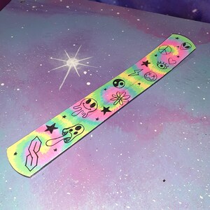 May include: A tie-dye incense stick holder with a variety of black and white illustrations, including a peace sign, a heart, an eye, a lightning bolt, a skull and crossbones, a yin yang symbol, a flower, a mushroom, a star, and an alien.