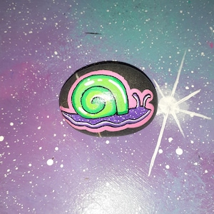May include: A hand-painted rock with a green snail design. The snail is painted on a black rock with pink and purple accents.