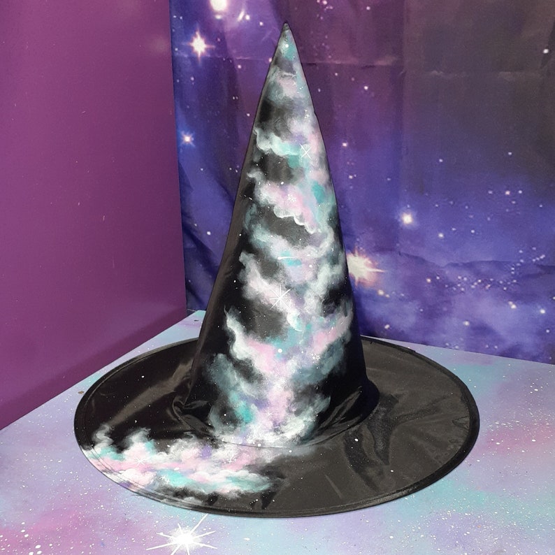 May include: A black witch hat with a galaxy print. The hat has a pointed cone and a wide brim. The galaxy print features swirling clouds of pink, blue, and white against a black background.