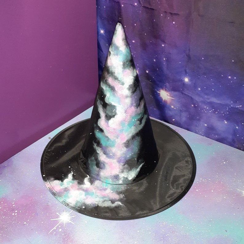 May include: A black witch hat with a galaxy print. The hat is painted with swirling clouds of pink, blue, and white, resembling a nebula. The hat is on a purple background with white stars.