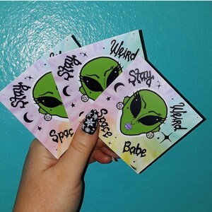 Op de afbeelding: Drie vierkante stickers met een groene alien met grote zwarte ogen en paarse lippen. De stickers hebben de woorden "Stay Weird Space Babe". De achtergrond is een tie-dye effect met roze, paarse en groene kleuren.