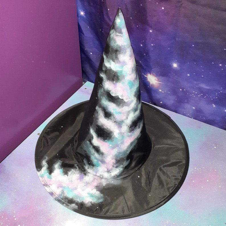 May include: A black witch hat with a wide brim and a tall pointed crown. The crown is painted with a galaxy design featuring swirling clouds of pink, blue, and white against a black background.