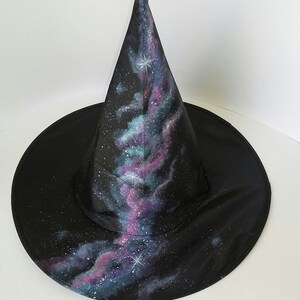 May include: A black witch hat with a galaxy print. The hat has a pointed cone and a wide brim. The galaxy print features shades of blue, purple, and pink with white stars.