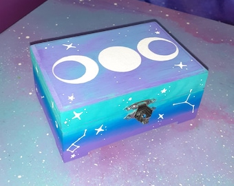 Puede incluir: Una caja rectangular de madera con un degradado pintado a mano en morado, azul y verde azulado. La tapa muestra tres fases lunares blancas, mientras que los lados están decorados con estrellas y constelaciones pintadas. Un cierre de metal plateado asegura la tapa.