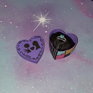 May include: A heart-shaped ring box with a black and white striped exterior and a purple interior holds a silver ring with a blue gemstone. The box is open next to a closed purple heart-shaped box with a silhouette design.