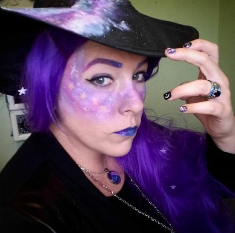 May include: A woman with purple hair and galaxy makeup is wearing a black witch hat with a galaxy design. She is also wearing a silver necklace with a blue pendant.