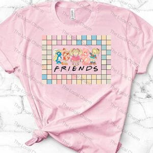 Puede incluir: Una camiseta rosa claro con un gráfico de personajes de dibujos animados y la palabra "FRIENDS" en negro. El gráfico está enmarcado por un borde a cuadros de colores pastel. La camiseta está anudada en la parte inferior.