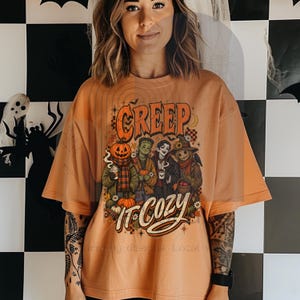 Könnte beinhalten: Ein orangefarbenes T-Shirt mit kurzen Ärmeln und einem Grafikdruck von vier Halloween-Figuren, darunter ein Kürbiskopf, ein grünes Monster, ein Vampir und eine Vogelscheuche, mit dem Text "CREEP IT COZY" in einer stilisierten orangefarbenen Schrift.