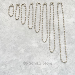 May include: A collection of silver-toned necklaces and bracelets with small, alternating silver and gold-colored beads. The jewelry is displayed on a light gray textured surface. The Radhika Store logo is visible in the bottom corner.
