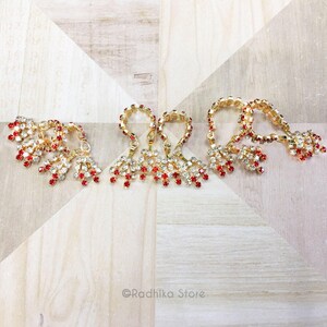 May include: Gold-toned jewelry with red and clear gemstones. The earrings are arranged on a light-colored wooden surface. The earrings have a loop design with dangling clusters of gemstones.