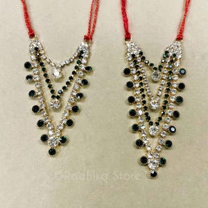 May include: Two ornate necklaces with gold-colored settings, clear and dark green gemstones, and red cords. The necklaces have a layered design with multiple strands of sparkling stones, and a teardrop shape.