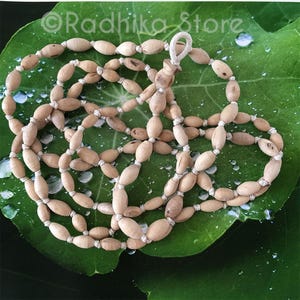 May include: A light brown wooden bead necklace with a knotted cord. The beads are oval-shaped and strung together with a white cord. The necklace is displayed on a green leaf with water droplets. The text "Radhika Store" is visible in the upper left corner.