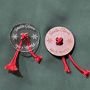 May include: Two round ornaments with red string attached. One ornament is clear with white snowflakes and the text "Santa Claus" and "North Pole". The other ornament is brown with gold glitter and the text "Santa Claus" and "North Pole".