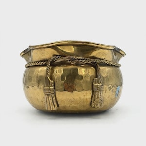 May include: A round, hammered brass bowl featuring a decorative rope-like trim encircling the upper section. The trim includes two hanging tassels made of the same brass material, positioned symmetrically on the front of the vessel.