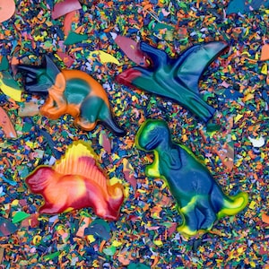 May include: Four dinosaur-shaped crayons in shades of orange, green, blue, and red, set against a background of crayon shavings. The crayons are a triceratops, a pterodactyl, a stegosaurus, and a T-Rex.