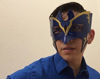 May include: A blue and gold masquerade mask with intricate details. The mask has a pointed top and a wide, open eye area. The mask is worn by a person with a blue shirt.