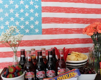May include: A Fourth of July party spread with red, white, and blue decorations. The spread includes a variety of snacks and drinks, including chips, cherries, and bottles of IBC root beer and cream soda. There are also sparklers and fireworks.