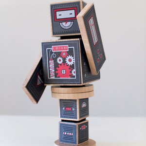 May include: A wooden robot toy with a gray and red color scheme. The robot is made of stacked blocks with different designs, including gears, a dial, and a red explosion. The robot is standing on a wooden base.