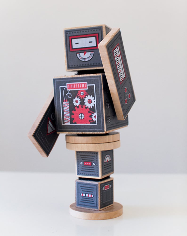 May include: A wooden robot toy with a gray and red color scheme. The robot is made of stacked blocks with different designs, including gears, a dial, and a red explosion. The robot is standing on a wooden base.