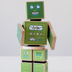 May include: A green wooden robot toy with a smiling face, blue eyes, and a control panel on its chest. The robot has movable arms and legs.