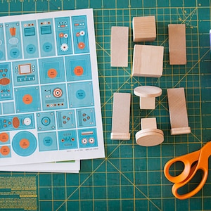 May include: A craft project with wooden blocks, a sheet of paper with colorful designs, a bottle of Mod Podge glue, and orange scissors on a green cutting mat.
