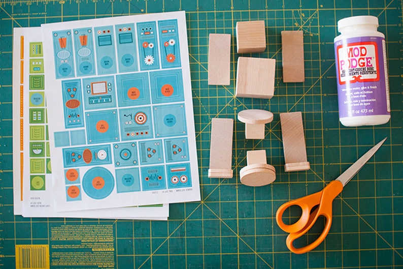May include: A craft project with wooden blocks, a sheet of paper with colorful designs, a bottle of Mod Podge glue, and orange scissors on a green cutting mat.