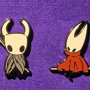 May include: Two enamel pins featuring characters from the video game Hollow Knight, next to a quarter for size comparison. One pin depicts a character with a white head, black eyes, and a gray cloak. The other pin shows a character in a red cloak.